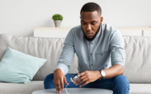 African Guy With Pulse Oximeter Clip On Hand Measuring Level Of Oxygen Saturation Sitting On Couch At Home