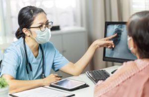 Asian female doctor in medical mask examining and pointing to radiological chest X-ray film on monitor computer while talking with Elderly woman patient at hospital room