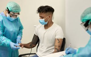 Medical professional in protective workwear placing pulse oxymeter on finger of seated man wearing surgical mask