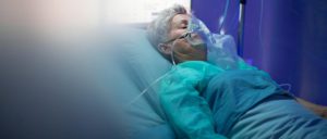 An older female patient lies in a hospital bed wearing an oxygen mask